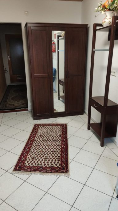 Holzschrank mit Spiegel in einem Raum, Teppich und Regal im Hintergrund.