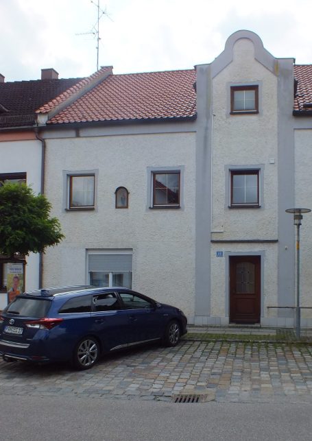 Helles Gebäude mit grauer Fassade, einem Auto davor und einem Baum an der Seite.